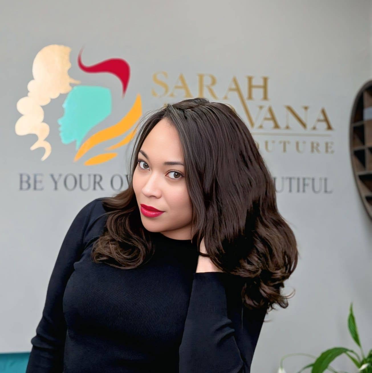 Sarah Afiovana in a black top, seated in front of the Hair Couture wall sign at the Matlock atelier.